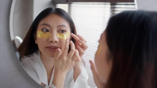Krásná asijská dáma použití pod očima skvrny při stání v blízkosti zrcadla v koupelně - Záběry, video