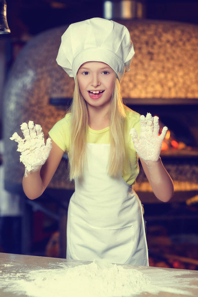 Egy kis vidám lány gyönyörű mosollyal, pékjelmezben, liszttel dolgozva, lisztben mutatja a kezét. - Fotó, kép