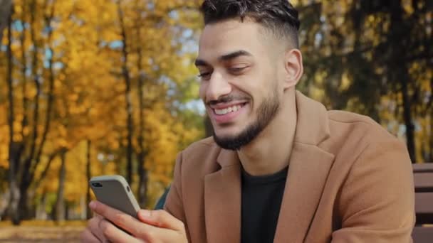 Primer plano joven hispano sentado en el banco en el parque de otoño barbudo hombre sosteniendo el teléfono móvil hace compras en línea sonriendo felizmente utilizando aplicación mirando en el teléfono inteligente al aire libre noticias de navegación en el dispositivo - Metraje, vídeo