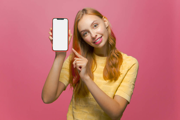 Cheerful young girl showing modern phone with blank mock-up screen. New entertaining or dating mobile app advertising - 写真・画像