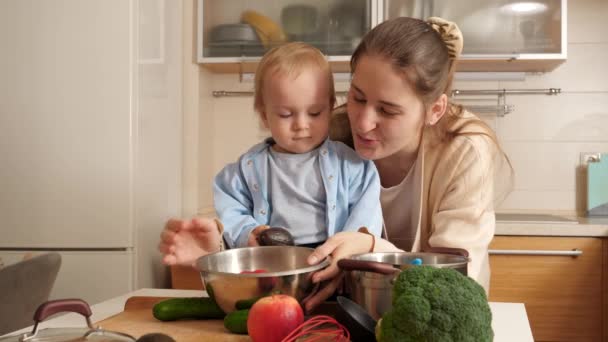 Νεαρή μητέρα δείχνει μαγειρικά σκεύη κουζίνας, σκεύη και συστατικά στο μικρό γιο της στην κουζίνα. Έννοια του μικρού σεφ, τα παιδιά μαγείρεμα των τροφίμων, καλές οικογενειακές στιγμές μαζί. - Πλάνα, βίντεο