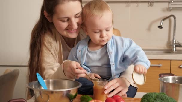 Usmívající se žena dává ovocné pyré svému malému synovi, který si hraje v kuchyni. Koncept malého šéfkuchaře, děti vařící jídlo, dobré společné rodinné chvíle. - Záběry, video
