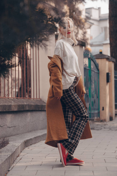 Fashionable beautiful young woman with blond hair in a stylish long coat, checkered pants, red shoes and glasses poses in the city streets. - Photo, Image