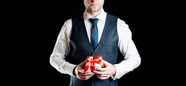 Geschenkmann. Glückliche junge Geschäftsmann mit Überraschung Geschenk-Box mit roter Schleife isoliert auf schwarzem Hintergrund. Schwarzer Freitag, Valentinstag Glückwunschbanner - Foto, Bild