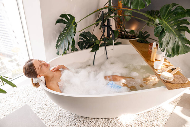 Relaxed young woman bathing with body care cosmetics on wooden shelf over modern white bubble filled bathtub at home - Foto, Imagem