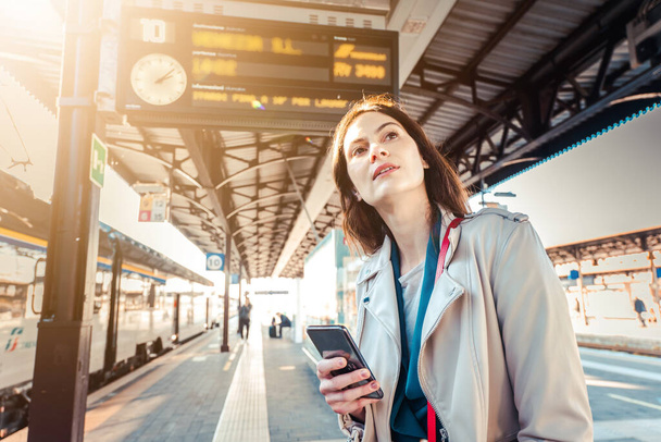 Jeune femme avec les heures de départ derrière elle attendant son train tout en tenant son téléphone portable - Femme regardant l'horloge dans la gare pendant que son train est retardé - Concept de transport et de vie urbaine - Photo, image