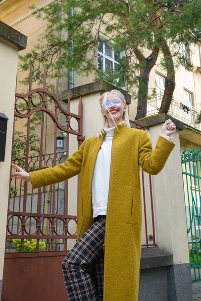 Fashionable beautiful young woman with blond hair in a stylish long coat, checkered pants and glasses poses in the city streets. - Fotoğraf, Görsel