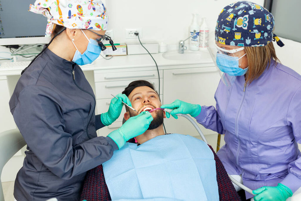Naishammaslääkäri tutkii miehen potilaan hammashoitolassa käyttäen ammattilaisen työkaluja ja henkilönsuojaimia. - Valokuva, kuva