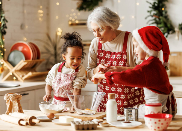 Happy multiracial kids help grandmother to cook Christmas cookies in kitchen during winter holidays - Fotó, kép