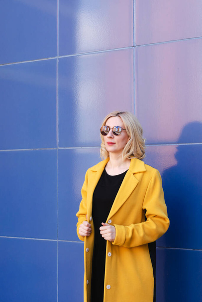 Young woman with blond hair in a bright yellow coat on a blue background on a city street. Urban style of modern woman - Photo, Image