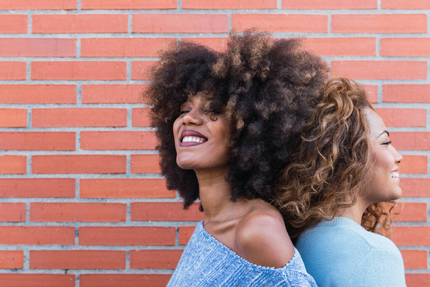 Oldalnézet mosolygós afro-amerikai nők hosszú afro haj és csillogás smink pózol téglafal. Szépség portré afrikai természetes lányok álló háttal másolás tér - Fotó, kép
