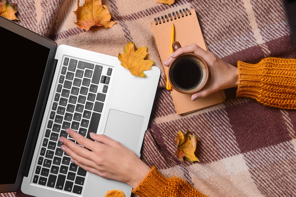 ラップトップ、コーヒー、ノートブック、秋の葉を持つ女性がチェックされたファブリックの背景に - 写真・画像