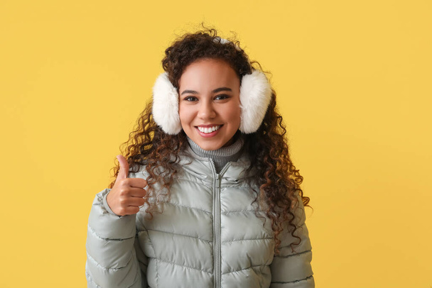 Pretty African-American woman in winter clothes showing thumb-up on color background - Foto, Bild