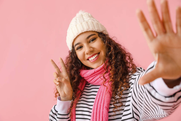 Pretty African-American woman in winter clothes showing victory gesture on color background - Fotó, kép