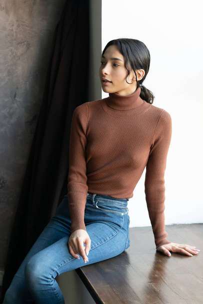 jovem bela mulher senta-se em jeans perto da janela no estúdio - Foto, Imagem