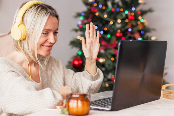 Boldog középkorú nő, fejhallgatóval és laptoppal a hátán egy karácsonyfa. Felnőtt hölgy kommunikál online, vagy néz egy webinar a karácsonyi ünnepek alatt - Fotó, kép