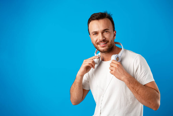 Mladý muž v ležérní bílé tričko se sluchátky proti modrému pozadí - Fotografie, Obrázek