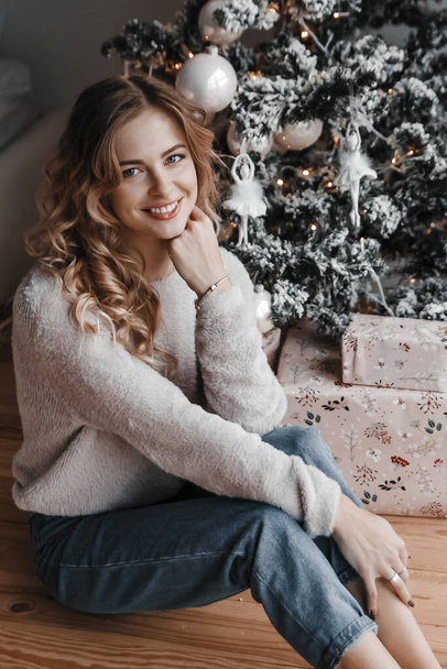 Woman is sitting by the christmas tree and presents. She is smil - Photo, image