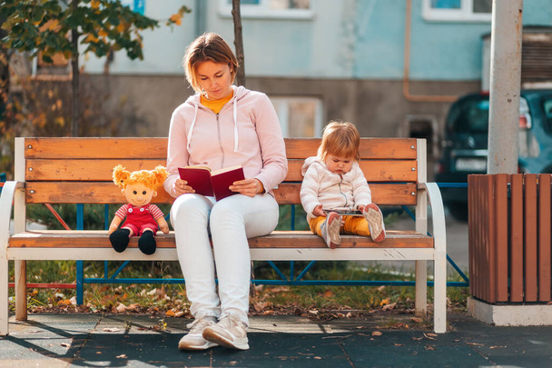 La joven madre está leyendo un libro sentado en un banco. Un niño está sentado cerca y viendo dibujos animados en un teléfono inteligente. - Foto, imagen
