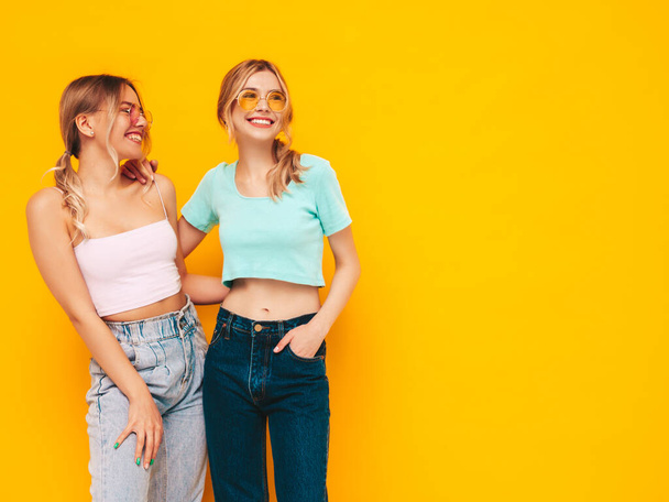 Two young beautiful smiling blond hipster female in trendy summer clothes. Sexy carefree women posing near yellow wall in studio. Positive models having fun. Cheerful and happy. In sunglasses - Valokuva, kuva