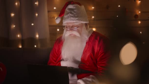 Santa está trabajando en casa con un portátil en Nochebuena. Luces de Navidad y decoración - Metraje, vídeo