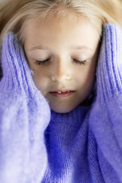 Cute little caucasian girl seven years old with blonde hair and closed eyes. Kid wearing stylish cozy knitted sweater violet color. Trendy color of the 2022 year very peri - 写真・画像