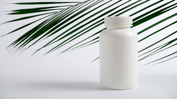 Weiße Plastikflasche für Vitamine, Kosmetika, Gesundheitsprodukte auf weißem Hintergrund mit grünen Palmblättern. - Foto, Bild