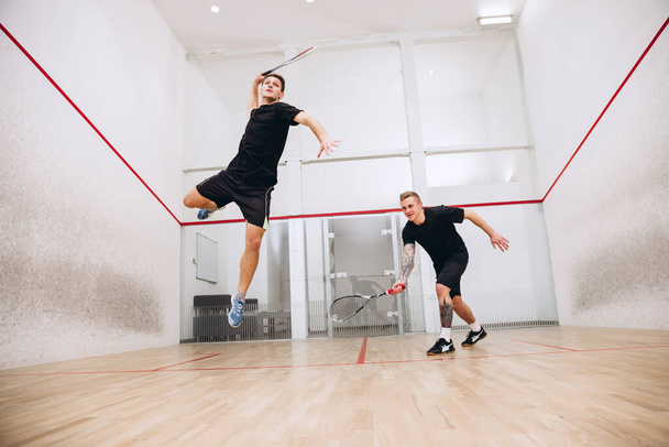 Full-délka portrét dvou mladých sportovních kluků trénink společně, hrát squash izolované nad sportovním studiem pozadí - Fotografie, Obrázek
