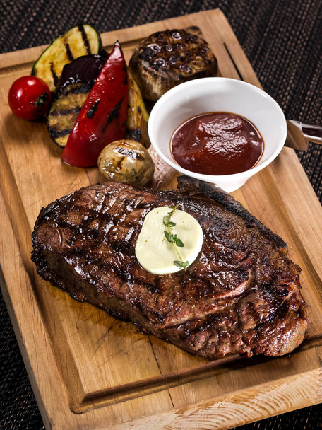 grilled beef angus steak with melted butter on a wooden cutting board served with grilled vegetables, close up photo on a dark background - Фото, зображення