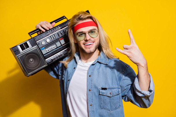 Photo of cute cool gentleman wear jeans shirt dark glasses listening boombox showing hard rock sign isolated yellow color background - 写真・画像