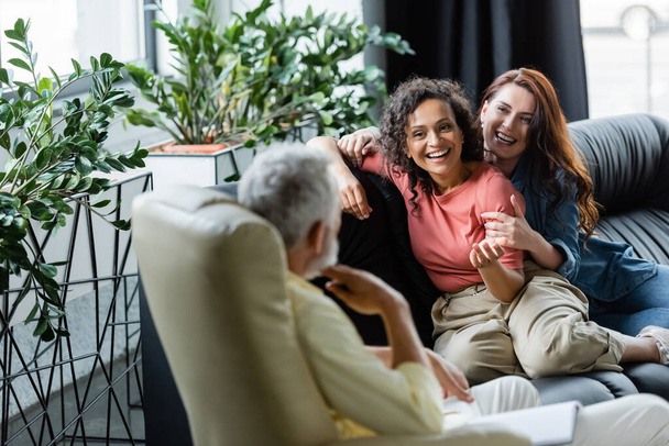 cheerful multiethnic lesbians sitting on couch near blurred psychologist - Photo, Image