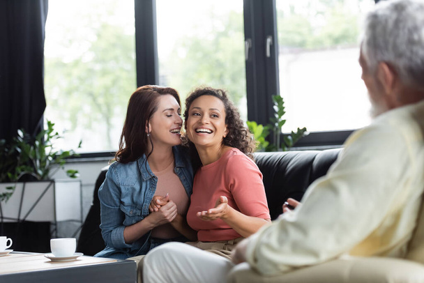 happy african american american woman talking to blurred psychologist while sitting on sofa with lesbian girlfriend - Valokuva, kuva