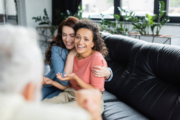 happy lesbian woman embracing african american girlfriend while sitting on sofa near psychologist on blurred foreground - 写真・画像