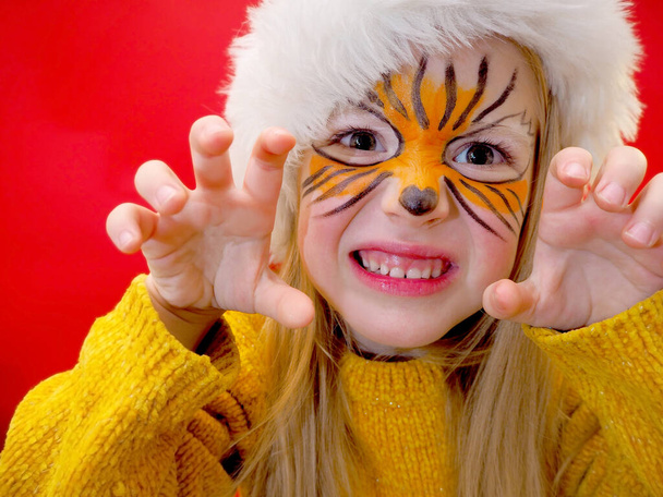 Child girl blonde with face painting in a tiger mask and Santa Claus hat growls and shows claws.  Year of the tiger.  Symbol of the year.  New Year. - Photo, Image