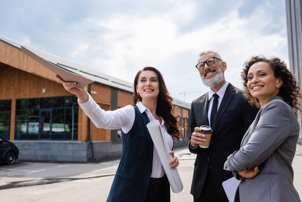 smiling interracial business colleagues looking away near real estate agent pointing with folder outdoors - Photo, Image