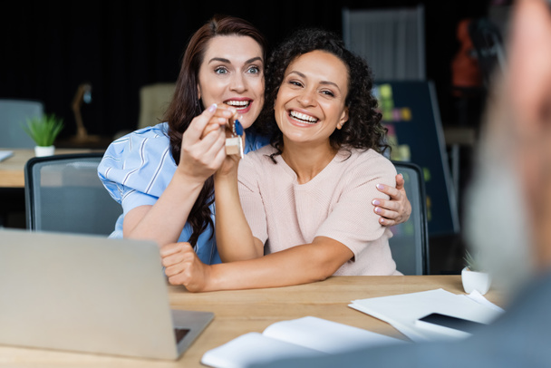 happy multiethnic lesbian couple holding key near blurred broker in real estate agency - Photo, Image