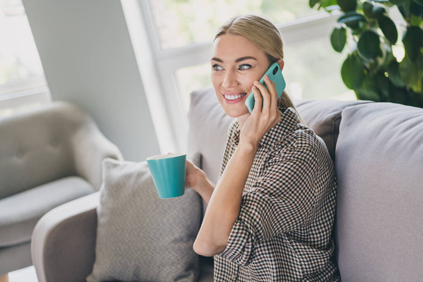 Photo of positive cheerful lady sit divan enjoy self isolation hold cappuccino mug call friend smartphone in apartment - Foto, Bild