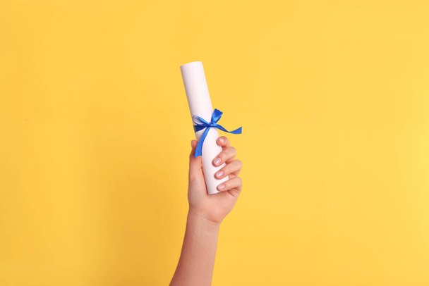 Student holding rolled diploma with blue ribbon on yellow background, closeup - Photo, image