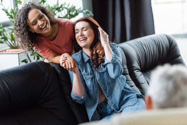 excited interracial lesbian couple holding hands and laughing near blurred psychologist during consultation - Photo, Image