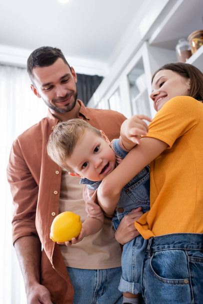 Smiling mother holding toddler son with lemon near husband at home  - Photo, Image