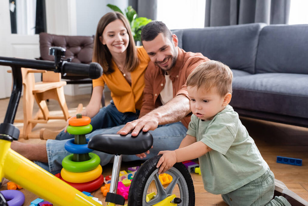 Dítě stojící poblíž kola a usmívající se rodiče v obývacím pokoji  - Fotografie, Obrázek