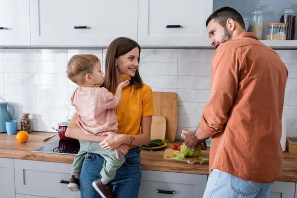 Усміхнена мати тримає дитину, коли чоловік ріже капусту на кухні
  - Фото, зображення