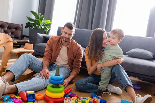 Mujer alegre abrazando hijo cerca de marido y juguetes en sala de estar  - Foto, Imagen
