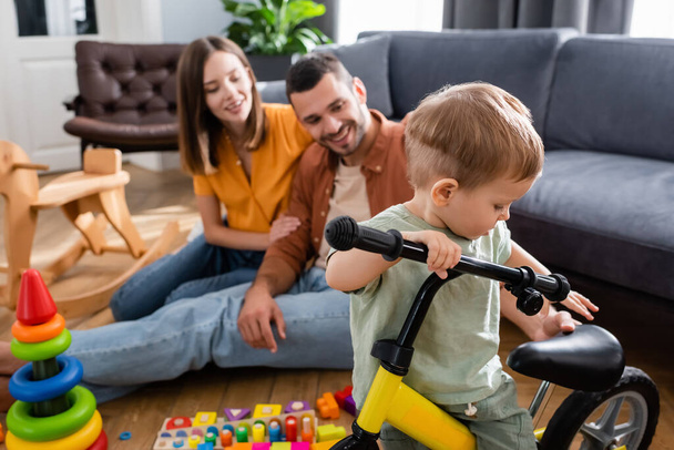 Toddler kid standing near bicycle and blurred parents in living room  - Foto, afbeelding