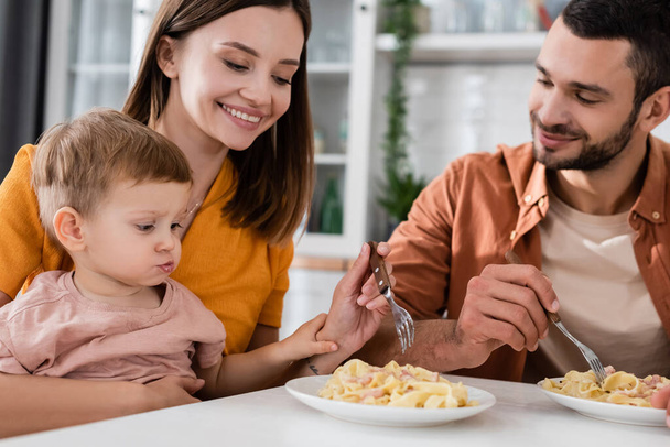 Smiling woman holding fork near pasta and family at home  - Foto, afbeelding