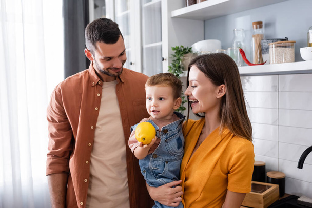 Positive parents looking at toddler son with lemon in kitchen  - Фото, зображення