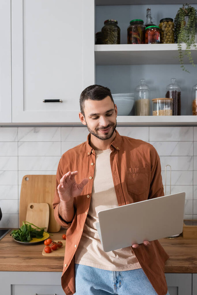 Smiling man having video call on laptop near vegetables in kitchen  - Photo, Image