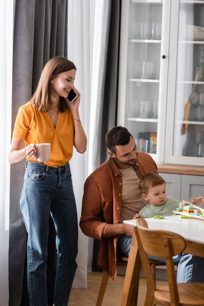 Nő csésze beszél okostelefonon, míg a férj és a fia játszik játék otthon  - Fotó, kép