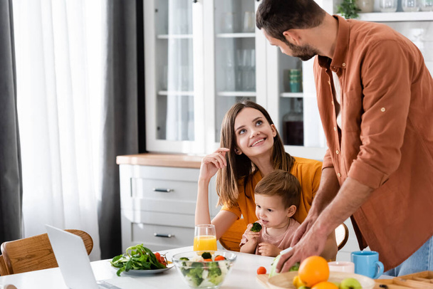 Smiling woman looking at husband cooking salad near kid and laptop in kitchen  - Foto, imagen