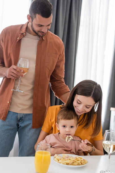 Man holding wine near wife feeding son with pasta at home  - Fotografie, Obrázek